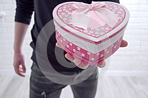 The guy holds in his hand a pink gift box in the form of a heart against the background of a white loft.