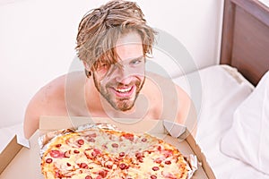 Guy holds pizza box sit bed in bedroom or hotel room. Student is at home on the bed in a bright apartment eating a tasty