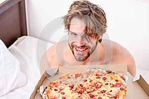 Guy holds pizza box sit bed in bedroom or hotel room. Student is at home on the bed in a bright apartment eating a tasty