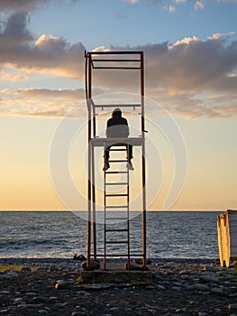 guy in a hood on a rescue tower. Silhouette of a young lonely man. Silhouette of a person looking out to sea at sunset.