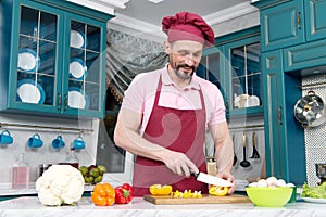 Guy with knife cutting orange pepper on table. Cook dressed in apron prepare dinner with paprika. Man in hat preparing vegetables.