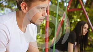 Guy looks towards the sad girl on a swing at the playground in the summer. Medium shot.