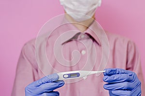 A guy in a mask and a medical glove holds a thermometer. Pink background.