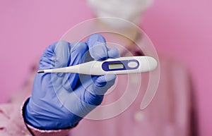 A guy in a mask and a medical glove holds a thermometer. Pink background.