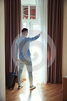 Guy opens curtained window standing with back in hotel room