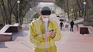 Guy with a phone standing in front of the camera on the stairs of the park he wearing protective mask while is the