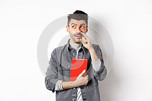 Guy picking nose and look aside at logo, holding red book or planner in hand, standing against white background