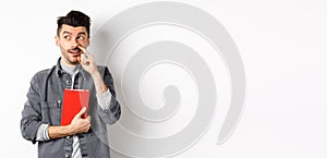 Guy picking nose and look aside at logo, holding red book or planner in hand, standing against white background