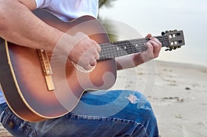 A guy plays an acoustic six-string guitar on a sandy beach by a body of water