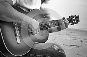 A guy plays an acoustic six-string guitar on a sandy beach near a body of water. Black-and-white image