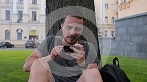 A guy plays a game on the phone while sitting under a tree in a park sitting under a tree