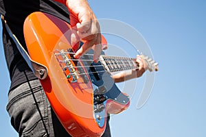 The guy plays an orange bass guitar against a cloudless blue sky