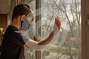 A guy in a protective mask sits at home on isolation and looks out the window while watching the street