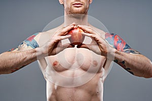 a guy with a pumped-up torso and a tattoo holds an apple in his hands on a gray background