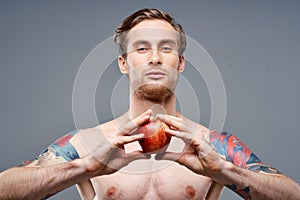 a guy with a pumped-up torso and a tattoo holds an apple in his hands on a gray background