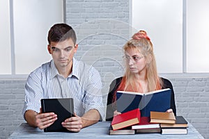 A guy is reading an e-book, and a girl is sitting at a pile of various books and gazing with charity at the guy.