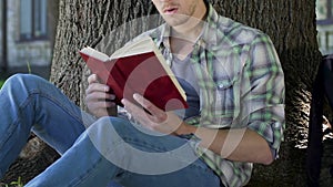 Guy reading gripping book under tree, engrossed into plot, avid book reader