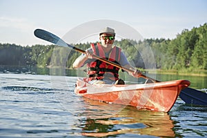 The guy is sailing on a red kayak, wearing a life jacket with glasses and a cap