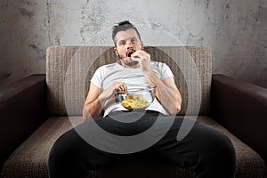 The guy in the shirt is lying on the couch, eating chips and watching a sports channel. The concept of laziness