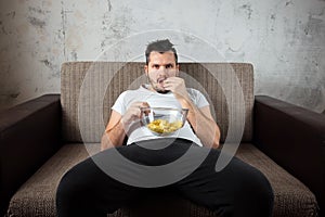 The guy in the shirt is lying on the couch, eating chips and watching a sports channel. The concept of laziness