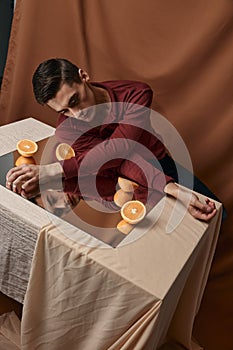A guy in a shirt sits at a table with a mirror and orange oranges fabric background