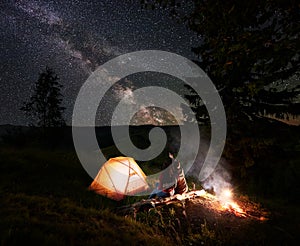 Guy shows girl up on evening starry sky at Milky way near tent and bonfire on background mountains