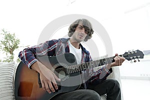 Guy sings a song and plays guitar sitting on the couch.