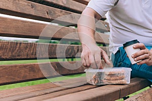 The guy sits on a bench and dines with a toast with a coffee in a glass. Fast food in the fresh air