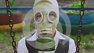 Guy sits on a swing between two rusty chains, adjusts soviet gas mask on face, look at the camera