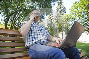 Guy sitting with laptop and talking on the phone in the park