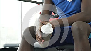 Guy sitting on training equipment in gym with protein drink, nutritive elements