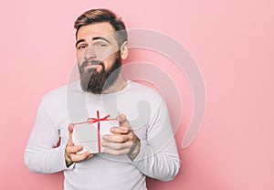 Guy is standing and holding a present. The box is white with red ribbon. Isolated on pink background.