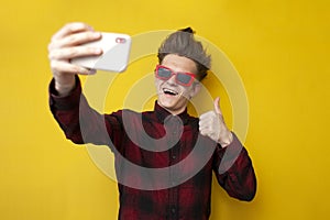 Guy takes selfie on smartphone and shows like on yellow isolated background, hipster in glasses takes pictures of himself and