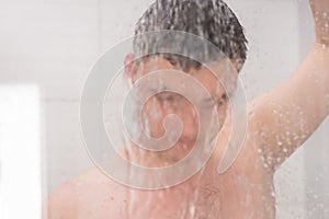 Guy taking a shower and holding shower head behind misted glass