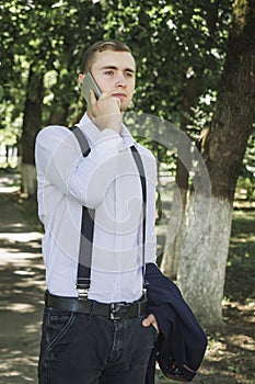 Guy talking on the phone. A young guy in jeans, a shirt and suspenders talking on the phone in the park.