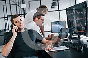 Guy talks by phone. Team of stockbrokers works in modern office with many display screens