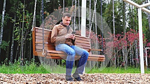 Guy texting on mobile phone in park. Young caucasian man relaxing on wooden swinging bench in autumn