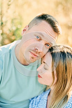 Guy touches girl head with his cheek while hugging her while sitting on the grass