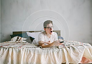 Guy using remote control while sitting on the bed in home. Happy attractive young man watching tv . Human emotions on