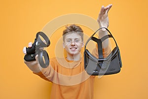 Guy in VR glasses on an orange background holds control bullets and offers to play, gamer shows a modern joystick, close-up
