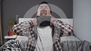 Guy wearing neck brace to support neck at home on couch. Caucasian man with neck injury uses collar support to relieve