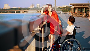 Guy On A Wheelchair And Girl With Red Hair Communicate On The Waterfront In The Summer Evening