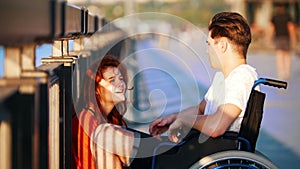 Guy On The Wheelchair Says Something To Red-Haired Girl And She Iistens Carefully, On Promenade In Summer Evening