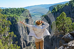 Guy in a white shirt and a brown hat, raising his hand, stands on a rock near a cliff in front of the grand canyon around the