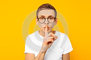 The guy in the white t-shirt in round transparent glasses says: Shhhh, shows silent gesture, on a yellow background