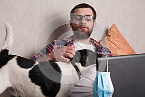 The guy works at home on freelance with his pet. The guy interacts with a dog on the couch with a medical mask