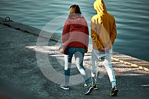 A guy in a yellow jacket and a girl in a red jacket walk down the berth near the water and look in front. Silence, peace and