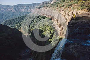 A guy in a yellow raincoat sits on the edge of a cliff near a waterfall on the background of a valley and mountains