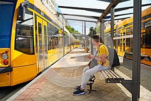 A guy in a yellow T-shirt is waiting for his tram at a public stop. There are yellow trams around him