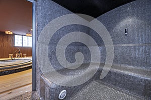 A gym sauna covered with gray tiles and steps to be used as a seat and exit to a circular jauzzy room